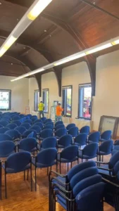 A large room with a vaulted wooden ceiling, hardwood floors, and rows of blue chairs. Two workers are on ladders, installing windows along one wall. Two stacks of empty blue chairs are in the foreground, and a large mirror leans against the wall in the background.