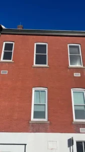 A vertical shot of a two-story red brick building with six white windows. The building has a white base and a dark trim under the roof. The sky above is a clear blue.