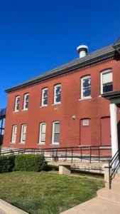 A side view of a two-story red brick building with multiple white windows. A red door is on the right, and a metal ramp with black railings leads up to a different red door on the left. A black staircase is visible on the far left of the building.