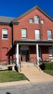 A tall red brick building with a two-story covered porch with a set of concrete stairs leading to the front door. A metal ramp with black railings is on the left, leading to a smaller red door. A black ladder is mounted to the wall above the front door.