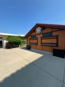 A wide-angle view of a brown, one-story building with a red roof. There are two windows with dark shutters on the front of the building, which has a circular sign above the windows. A concrete patio is in front of the building, and a white, ivy-covered building is in the background. The sky is clear and blue.
