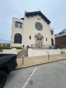 A straight-on view of the front of a large, two-story building. The building is made of cream-colored stucco with a dark, tiled roof. The front entrance has an ornate stone archway, and there are several large windows. Concrete steps lead up to the building from the street level. A black pickup truck is parked on the left side of the frame.