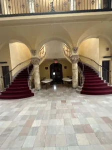 A vertical, straight-on view of the main entrance to a large, elegant banquet hall. Two curved staircases with red runners lead up to an upper balcony with a black, ornate railing. The room has a tiled floor, pillars, and arches, and there are two small, round tables at the bottom of the stairs.