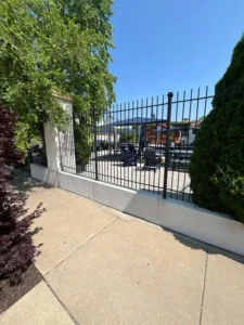 A horizontal, eye-level shot of a modern outdoor patio seen through a black metal fence. The patio has several gray chairs and tables under a blue canopy. The fence is on top of a short, white wall. A sidewalk is visible in the foreground, with trees and bushes on either side.