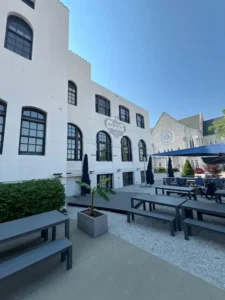 A view of a modern outdoor patio behind a white, multi-story building. The patio has several gray picnic tables and benches, as well as blue umbrellas and chairs. The patio is covered in light-colored gravel. A church with a large, circular stained-glass window is visible on the right side of the frame.