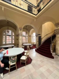 A vertical shot of a large, elegant banquet hall. There is a grand, curved staircase with a red runner on the right side of the frame, and a balcony with an ornate, black railing above it. The room is decorated with arches and pillars, and there are several round tables with gold chairs arranged in the center. A person is sitting at one of the tables on the left.