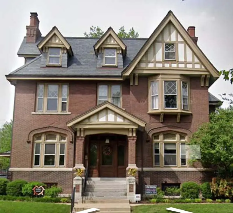 A straight-on view of the front of a large, two-story, red brick house with a steep slate roof. The house has a Tudor-style design with white and brown trim. There are several windows on the front of the house, including a large bay window on the right. A set of concrete steps leads up to a covered front door with a wreath on it. The yard is well-maintained, with green grass and manicured bushes.